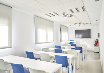 Sala de reuniones equipada con mesas y sillas orientadas hacia un estante y una pantalla para presentaciones o visualización de contenido